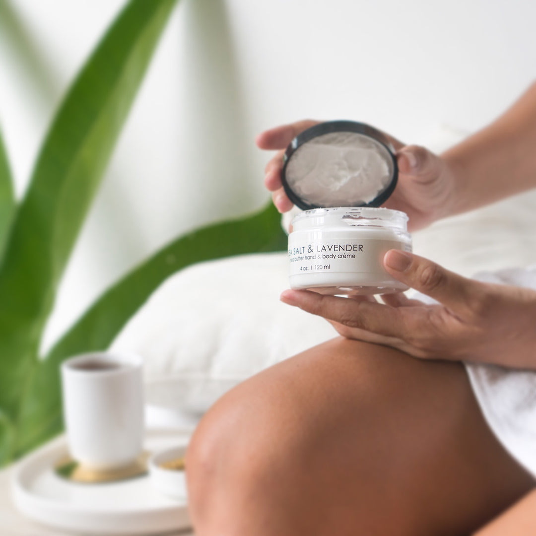 A person with bare legs, seated on a light-colored surface, holds an open jar of "SALT &amp; LAVENDER" body crème. A white tray beside them holds a cup, bowl, and other small items, including shea butter hand cream.