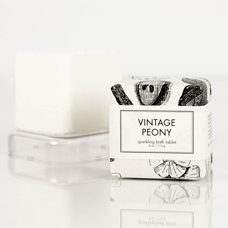 A white Formulary55 "vintage peony" Sparkling Bath Bomb Tablet rests on a clear plastic holder. The packaging, decorated with black and white illustrated peonies, reads "sparkling bath tablet" and specifies the weight as 4 oz/113g against a plain white background.