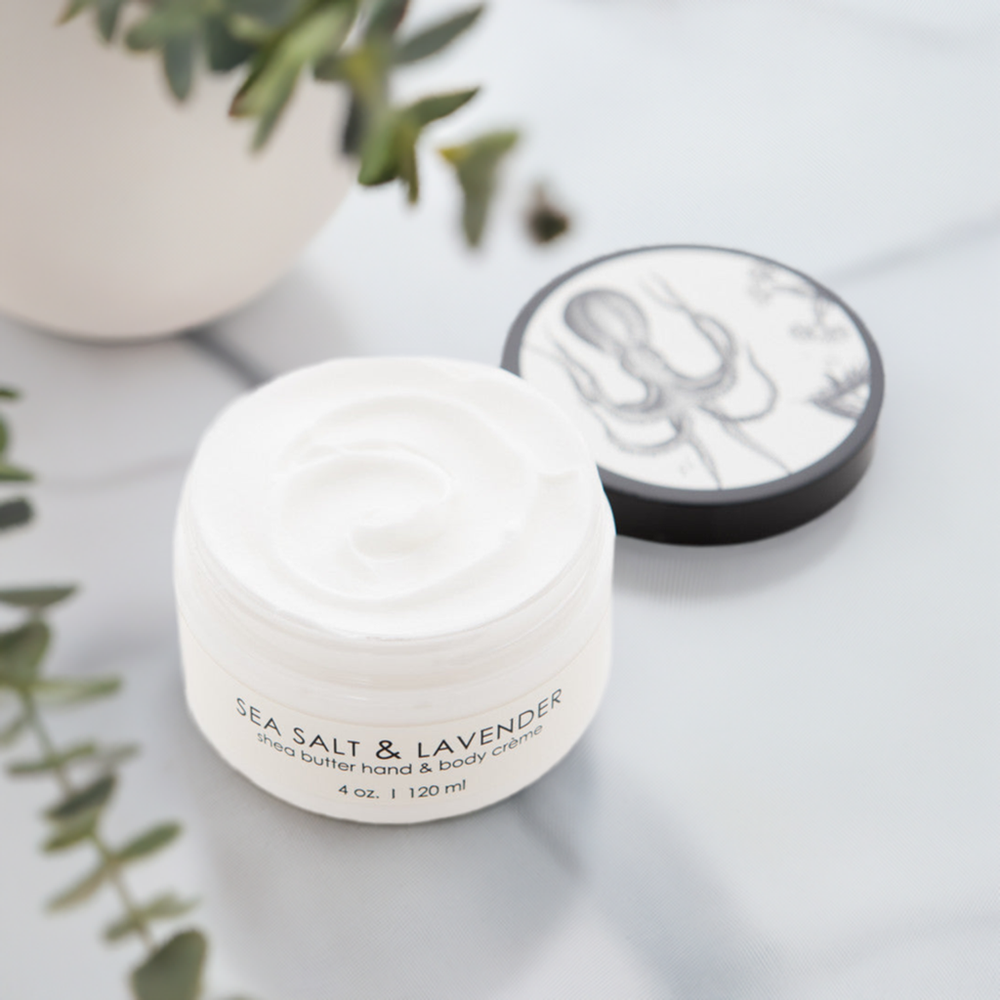 A small, white jar of Sea Salt & Lavender shea butter hand and body cream with its lid off. The jar sits on a light-colored surface surrounded by greenery. The black lid, featuring an illustration, is placed to the right of the jar.