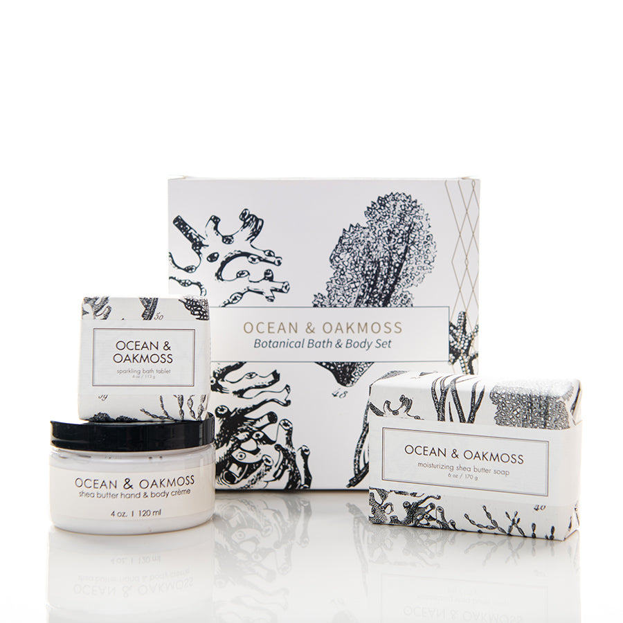 A neatly arranged display of Ocean & Oakmoss Botanical Bath & Body Set featuring three products: a jar of shea butter hand & body crème, a boxed moisturizing shea butter soap, and a decorative large box with matching botanical artwork.