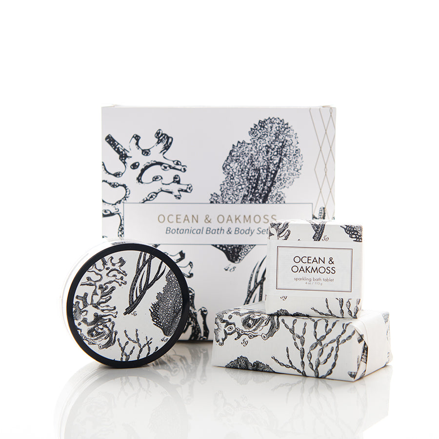 A botanical bath and body set labeled "Ocean & Oakmoss" consists of a boxed item, a circular container, and a bar of soap in white packaging adorned with black seaweed and coral illustrations. The items are neatly arranged against a white background.