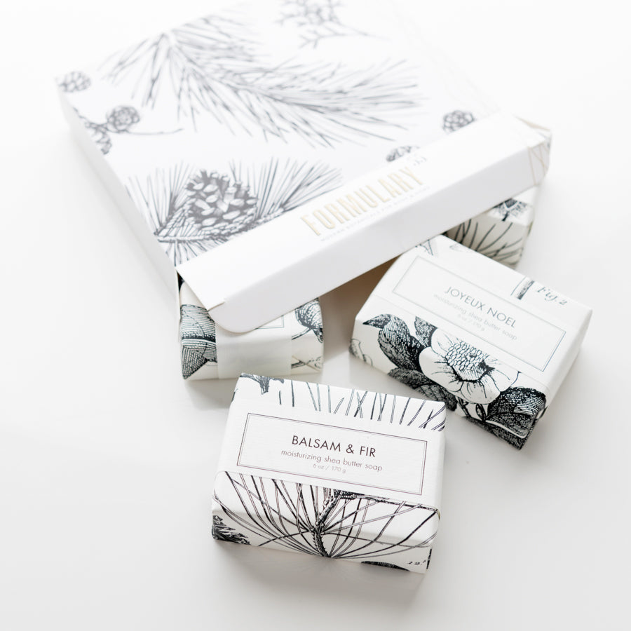 A set of four Shea Butter Bath Bars with different scents, including Balsam & Fir, Spiced Orange & Cedar, Sugared Plum & Fig, and Joyeux Noel, each wrapped in individual white packaging with botanical designs. The set is presented with a larger box that has a floral design.