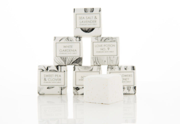 A white pyramid of small boxed soaps with labels reading "Sea Salt & Lavender," "White Gardena," "Love Potion No. 9," "Sweet Pea & Clover," "Southern Flowers Honey," and one unwrapped soap square in front, all against a white background.