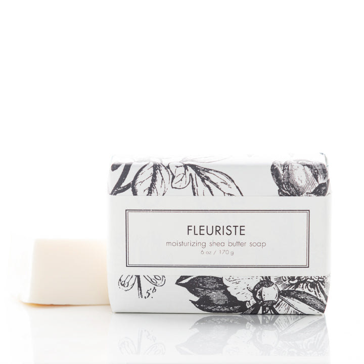 A bar of moisturizing shea butter soap labeled "Fleuriste" is shown. The soap is partially unwrapped, revealing a white, smooth bar. The packaging features an elegant black floral design on a white background. The soap weighs 6 oz (170 g).