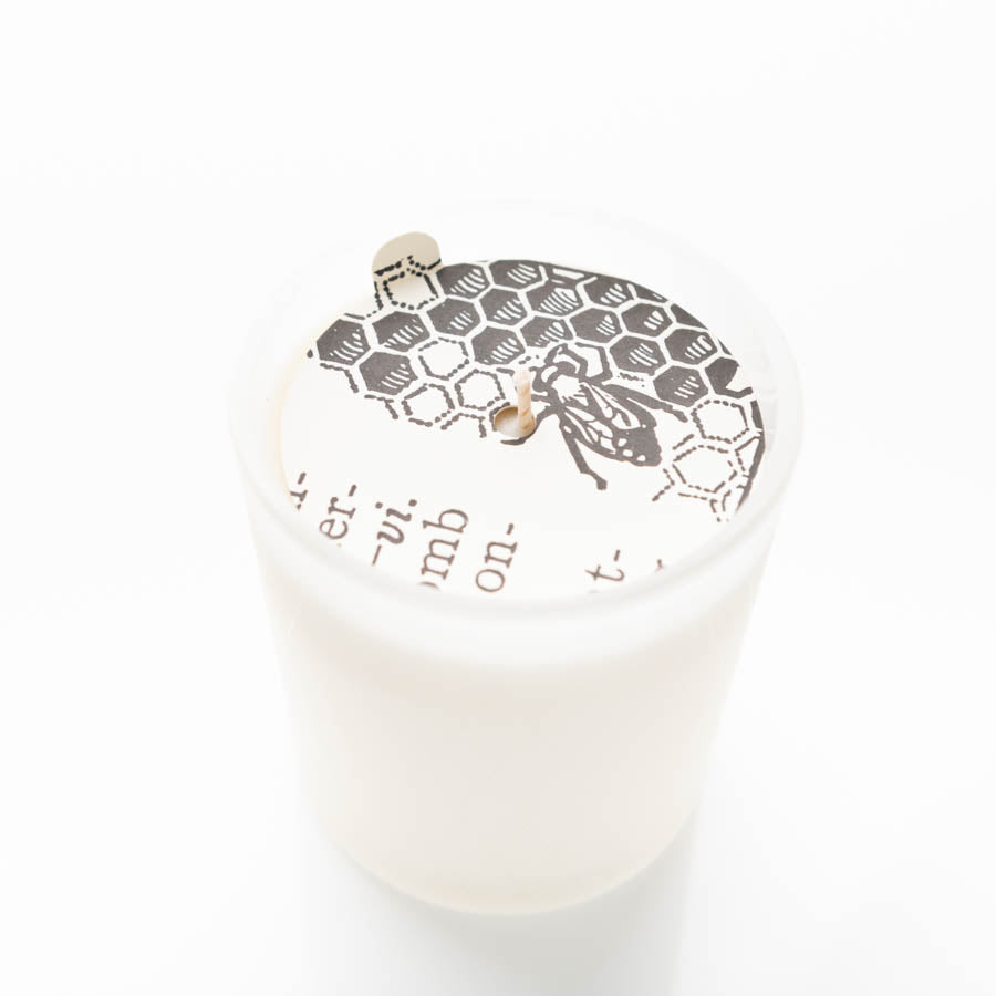 A white candle in a translucent glass container, featuring a black and white honeycomb pattern with a bee design on the surface. A small wooden wick is protruding from the center, surrounded by the design on the top. The background is plain white.