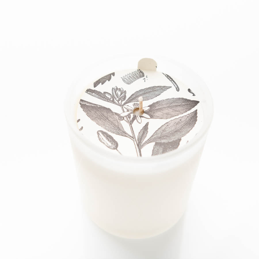 A white candle in a frosted glass holder. The top of the candle features a detailed black-and-white illustration of leaves, flowers, and bees. A single wick is centered in the middle of the illustration. The background is plain white.