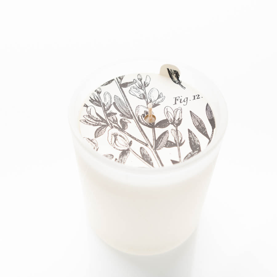 A white candle in a frosted glass holder features intricate black botanical illustrations on its surface. The top of the candle is adorned with floral imagery and labeled "Fig. 12." The wick is centrally placed. The background is plain white.