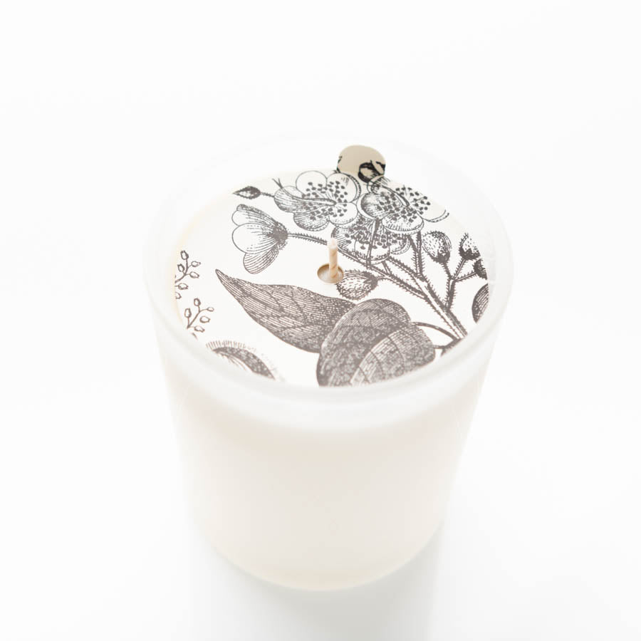 A white candle in a frosted glass holder, featuring an intricate black and white floral design on the top surface around the wick. The candle wick is in the center, surrounded by the detailed botanical illustration. The background is plain white.