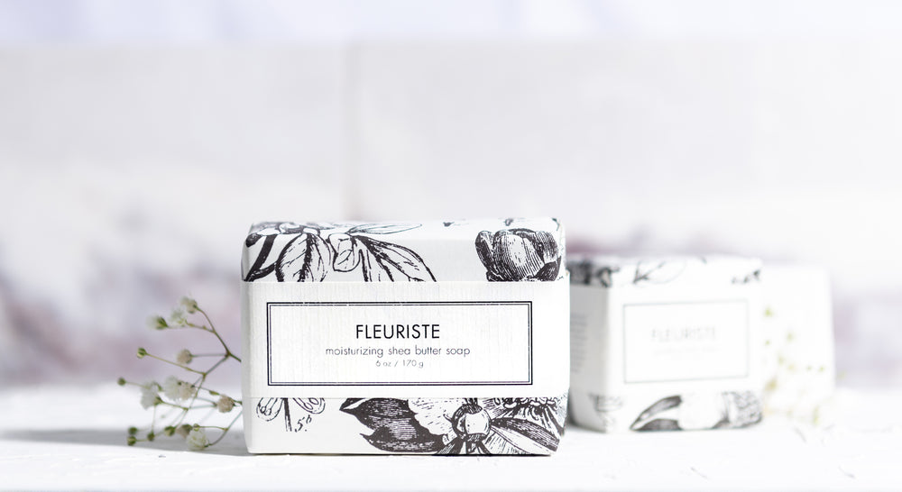 A bar of Fleuriste moisturizing shea butter soap is wrapped in white paper with black and white floral illustrations and is placed on a white surface. A smaller wrapped soap bar with the same design is partially visible in the background.