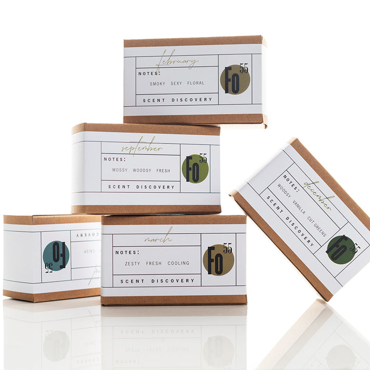 Five stacked boxes of scented products, each labeled by month with different scent profiles. February's "smoky, sexy, floral," March's "zesty, fresh, cooling," September's "mossy, woody, fresh." The minimalist packaging features green, gold, and gray accents.