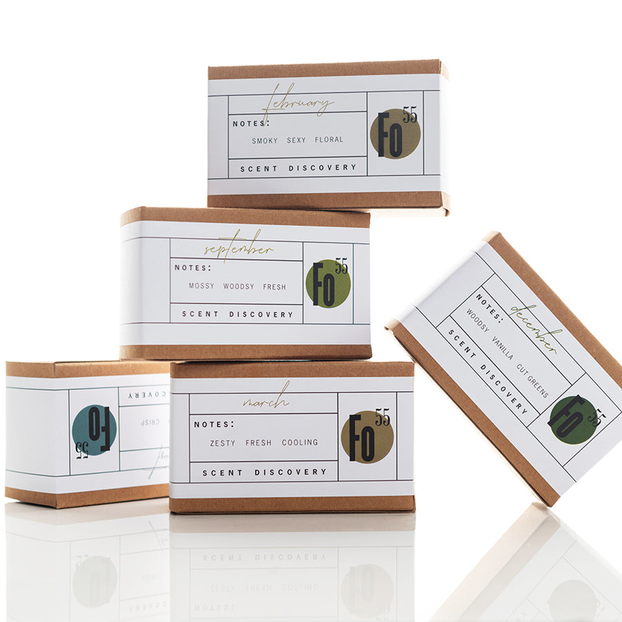 Five stacked boxes of scented products, each labeled by month with different scent profiles. February's "smoky, sexy, floral," March's "zesty, fresh, cooling," September's "mossy, woody, fresh." The minimalist packaging features green, gold, and gray accents.