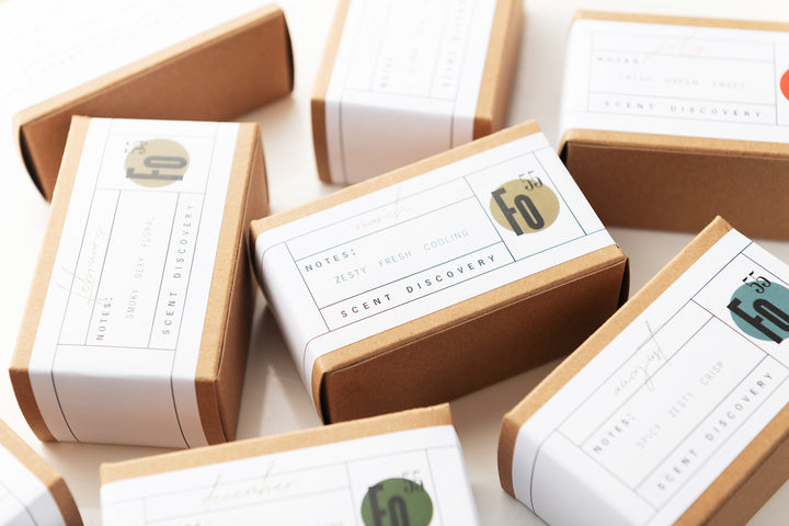 An arrangement of brown cardboard boxes with white labels. Each label has text describing the scent notes such as "Zesty", "Fresh", and "Cooling". The text "Scent Discovery" is also visible on the labels. The boxes are scattered on a light surface.