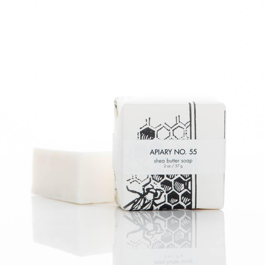 A bar of Apiary No. 55 shea butter soap is shown, with one bar partially unwrapped and another wrapped in packaging featuring a black and white design with honeycomb and bee illustrations. The label reads "Apiary No. 55 shea butter soap.