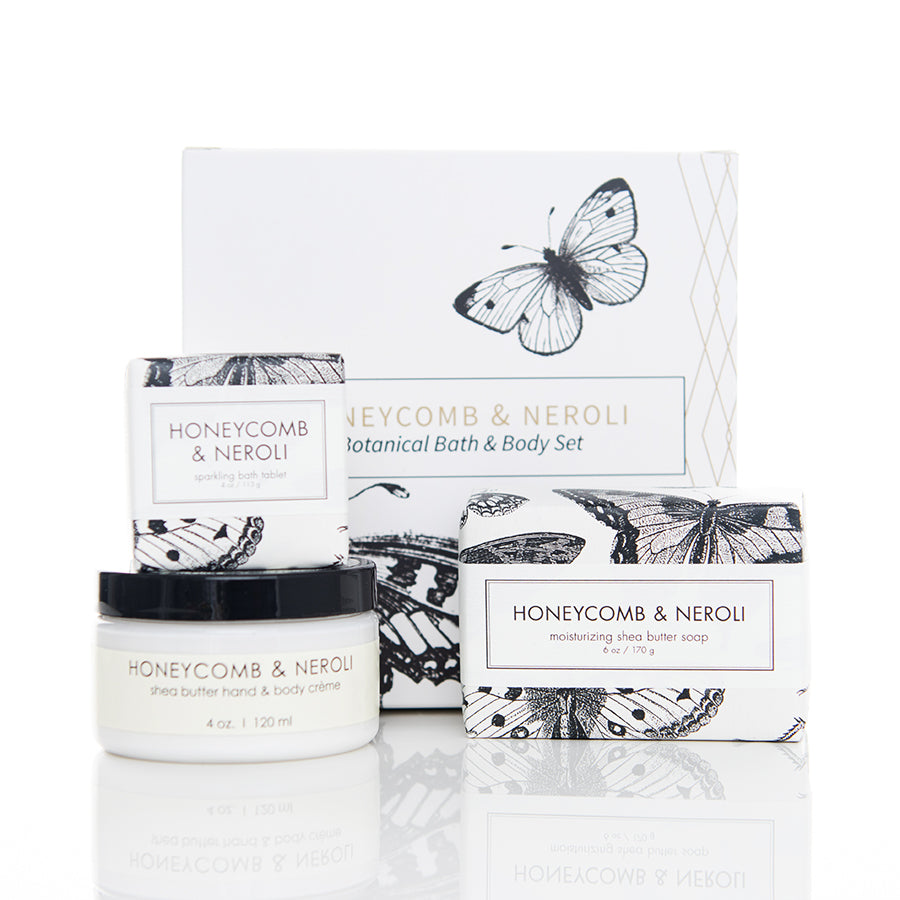 A white box with butterfly illustrations contains a Honeycomb & Neroli botanical bath and body set. The set includes three products: shea butter hand & body crème in a jar, a moisturizing shea butter soap, and a sparkle body balm, all in consistent packaging.
