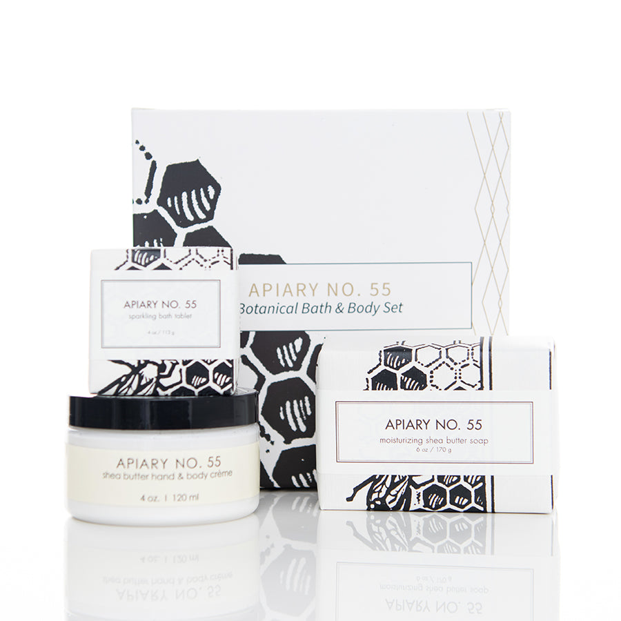 A botanical bath and body set featuring a shea butter hand and body crème, a moisturizing shea butter soap, and a sparkling bath tablet. The packaging is white with black geometric designs inspired by honeycombs. The product is labeled "Apiary No. 55.