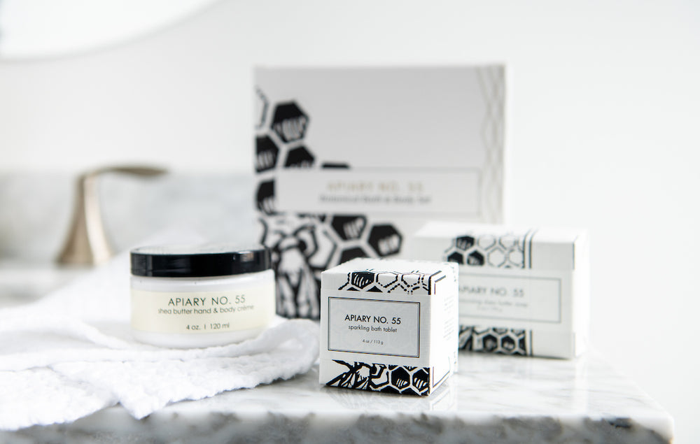 A bathroom countertop displaying Apiary No. 55 products, including a jar of shea butter hand and body cream and two boxes of sparkling bath tabs. The products feature a white and black minimalist design with honeycomb patterns.