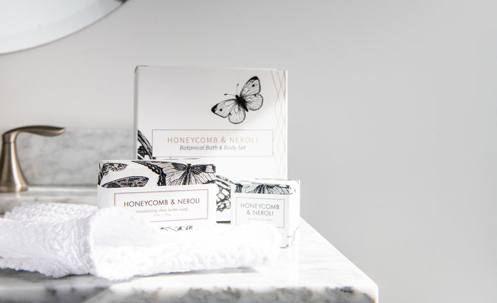 A luxurious bathroom set including a box labeled "Honeycomb & Neroli Botanical Bath & Body Set," a bar of moisturizing shea butter soap, and a lotion. The products feature elegant black and white illustrations of butterflies. A white towel is placed nearby.
