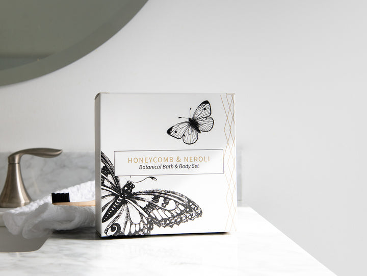 A botanical bath and body set box with "Honeycomb & Neroli" written on it rests on a white marble countertop. The box is decorated with intricate black-and-white illustrations of butterflies. A sink faucet and a rounded mirror are partially visible in the background.