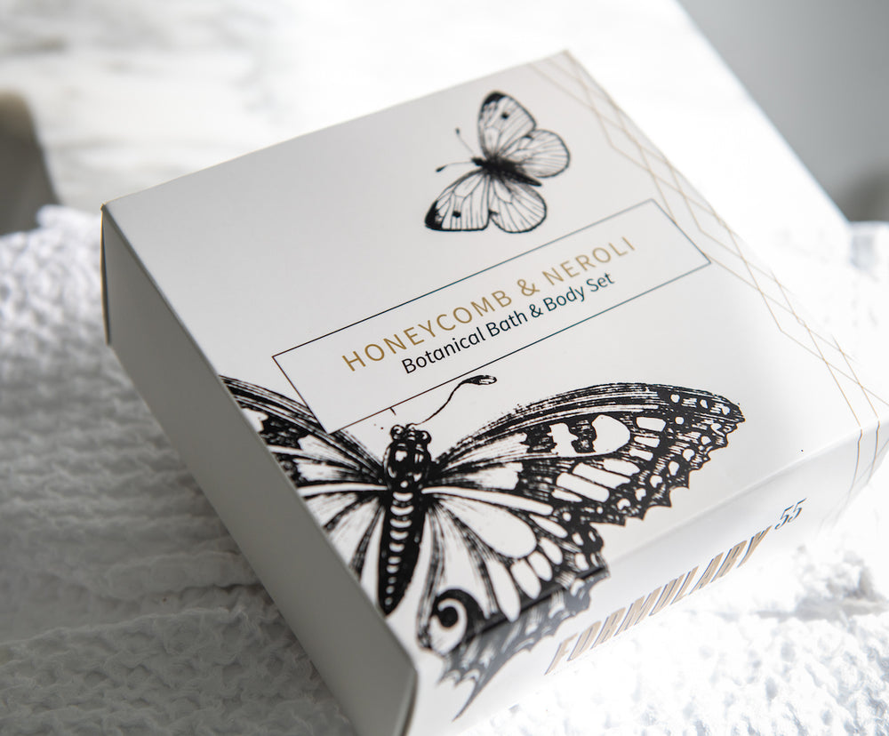 A white box with black illustrations of butterflies contains a "Honeycomb & Neroli Botanical Bath & Body Set." The box is resting on a textured white surface. The packaging design is elegant and minimalist.