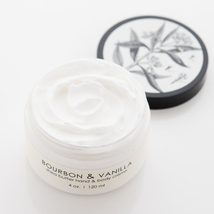 Image of an open jar of shea butter hand and body crème with the label "Bourbon & Vanilla" visible on the front. The jar is 4 oz. / 120 ml. The lid, placed beside the jar, features a black and white botanical illustration.