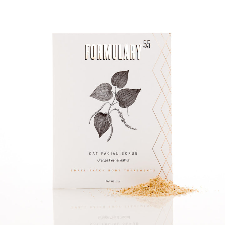 A white box of Formulary 55 Oat Facial Scrub with Orange Peel & Walnut. The front features a botanical illustration and gold geometric accents. A small pile of the scrub, beige in color, is scattered in front of the box. Text indicates it is a small batch body treatment.
