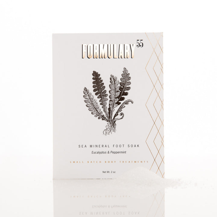 A product packaging for Formulary 55 Sea Mineral Foot Soak. The white box features an illustration of a fern in the center. Text indicates the eucalyptus and peppermint scented foot soak weighs 2 ounces. There is gold geometric design on the right side.