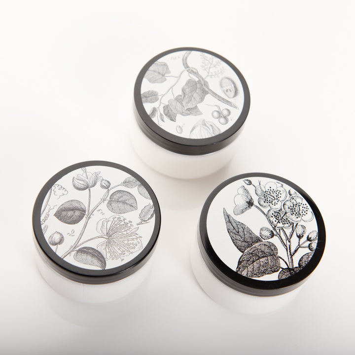 Three white circular jars with black lids are arranged on a white surface. Each lid features a different black-and-white botanical illustration, consisting of various flowers, leaves, and plant elements.