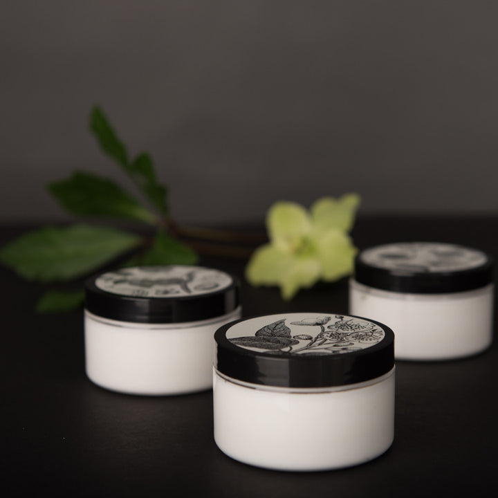 Three white jars with black lids, featuring floral designs, are arranged on a dark surface. A green leafy branch with a pale yellow flower is blurred in the background. The overall aesthetic is minimalistic and elegant.