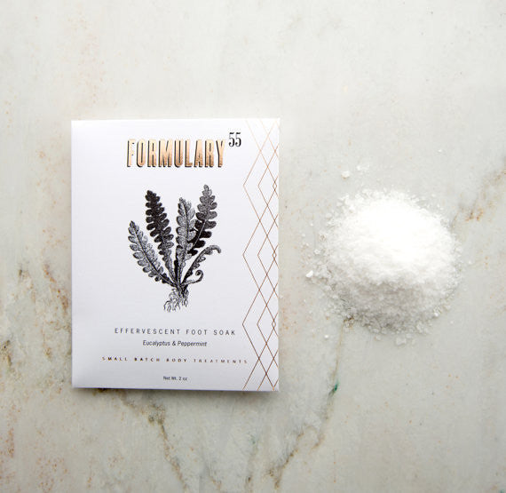 A white packet labeled "FORMULARY 55 Effervescent Foot Soak Eucalyptus & Peppermint" with an illustration of fern leaves is placed on a marble surface. Next to the packet is a small pile of white crystalline foot soak powder.