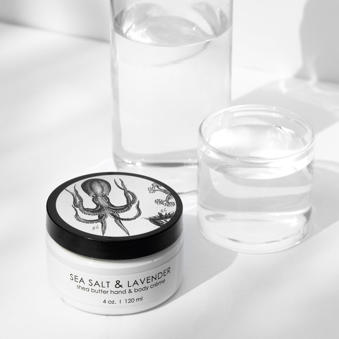 A jar of Sea Salt & Lavender shea butter hand and body crème is positioned in front of clear water glasses against a white background. The jar features an illustration of an octopus on its black lid.