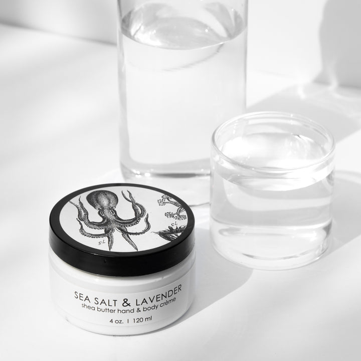 A jar of Sea Salt & Lavender shea butter hand and body crème is positioned in front of clear water glasses against a white background. The jar features an illustration of an octopus on its black lid.