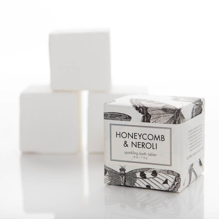 A cube-shaped package labeled "HONEYCOMB & NEROLI sparkling bath tablet, 4 oz/113g" with black and white butterfly illustrations. Three fuzzy white cubes are in the blurred background. The setup is on a white reflective surface.