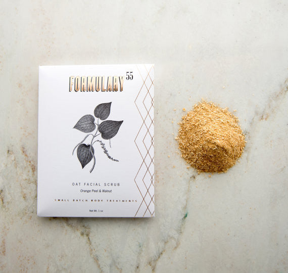 A package of Formulary 55 oat facial scrub labeled "Oat Facial Scrub: Orange Peel & Walnut" is placed on a marble surface next to a small pile of the granular scrub product. The package design features an illustration of plant leaves.
