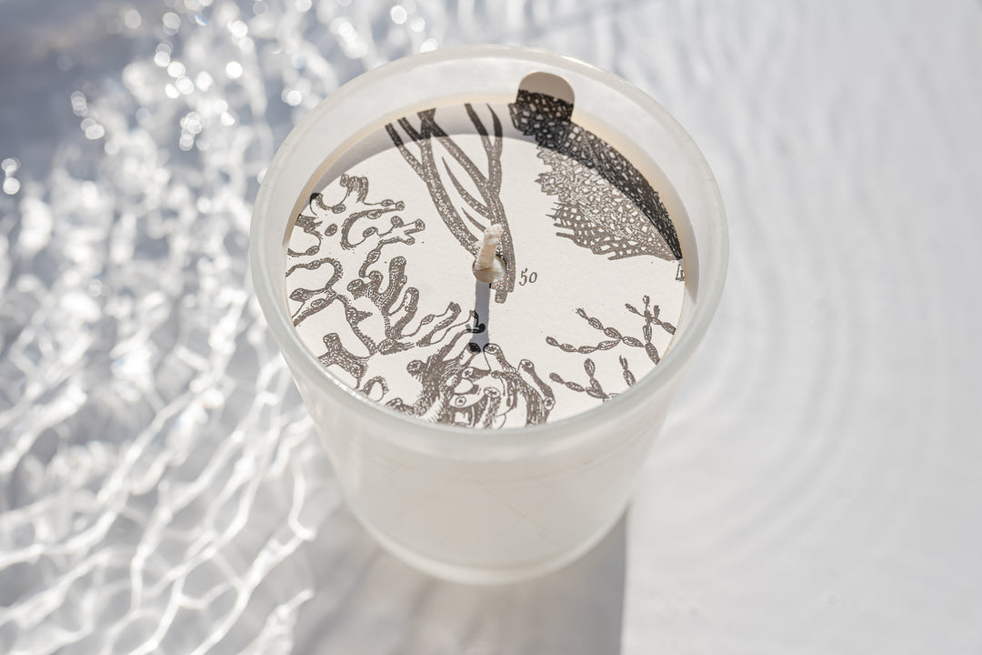 Close-up of a white candle in a clear glass holder, the surface of the candle features intricate black floral and botanical designs. The candle is placed on a textured white surface with shimmering light reflections surrounding it.